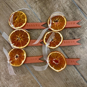 Könnte beinhalten: Getrocknete Orangenscheiben, gebunden mit weißen Bändern, angeordnet mit terrakottafarbenen Namensschildern. Die Schilder tragen Namen wie "Callum", "Mum", "Becky", "Dad" und "Al".