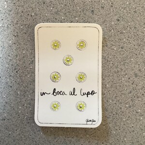 May include: A white card with seven yellow flowers and the text "in bocca al lupo" in black cursive writing. The card has a black border and a small signature in the bottom right corner.