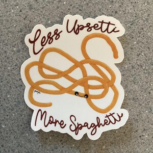May include: A white sticker with a brown outline and the text "Less Upsetti More Spaghetti" in brown. A cartoon spaghetti noodle with a face is drawn in the center of the sticker.