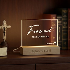 May include: A lighted acrylic sign with the words "Fear not, for I am with you" and the date "Baptism 26.10.2025" engraved on a wooden base. The sign is on a wooden shelf with books and a crucifix.