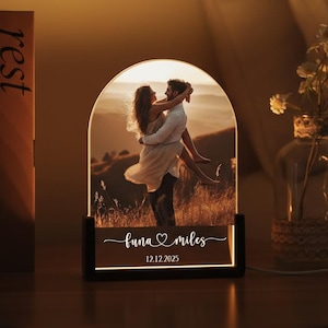 May include: An illuminated LED lamp with a photograph of a couple embracing, set within an arched frame. The lamp has a black base. The text "luna miles" and the date "12.12.2025" are displayed. The lamp is on a wooden surface, with books and a vase nearby.