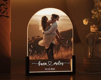 May include: A personalized LED lamp featuring a couple embracing, set against a scenic backdrop. The lamp has a black base and an arched frame. The text "luna miles" and the date "12.12.2025" are illuminated on the base. The lamp is on a wooden surface.