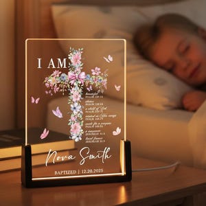 Può includere: Una luce notturna a LED personalizzata con un design a croce floreale. La luce presenta il testo "I AM", un nome e una data di battesimo. La croce è adornata con fiori rosa e viola e farfalle. La luce poggia su una base nera.