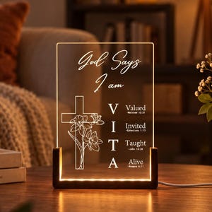 Può includere: Una targa luminosa rettangolare in acrilico con base in legno scuro. La targa presenta il testo "God Says I am" sopra una "VITA" verticale con le parole "Valued", "Invited", "Taught", "Alive" e versetti biblici, accanto a un grafico con croce e fiori.