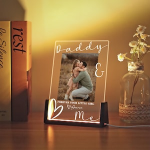 Può includere: Una cornice fotografica in acrilico illuminata con le parole "Daddy & Me" e una foto di un padre e sua figlia. La cornice ha una base nera ed è illuminata da una calda luce. È visibile anche il testo "Forever Your Little Girl".