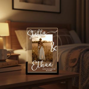 Peut inclure: Lampe de chevet LED personnalisée avec une photo d'un couple. Les noms "Stella & Ethan" sont illuminés en écriture cursive, avec le texte "LOVE YOU FOREVER 24.12.2025". La lampe a une base noire et un panneau acrylique transparent.