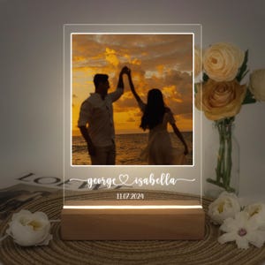 Może przedstawiać: Spersonalizowany akrylowy znak świetlny ze zdjęciem pary tańczącej na plaży o zachodzie słońca. Znak ma wygrawerowane imiona "george ♥ isabella" i datę "11.07.2024".