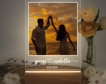 Op de afbeelding: Een gepersonaliseerd acryl lichtbord met een foto van een stel dat op een strand bij zonsondergang danst. Het bord heeft de namen "george ♥ isabella" en de datum "11.07.2024" gegraveerd.