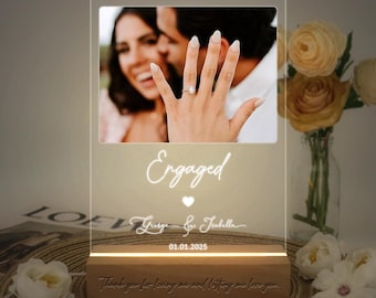 Op de afbeelding: Een wit LED-licht met een foto van een verloofd stel. Het licht heeft de tekst "Engaged" en "George & Isabella" met de datum "01.01.2025" onder de foto. De basis van het licht is van hout en heeft de tekst "Thank you for loving me and letting me love you."