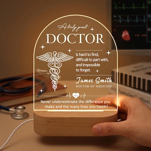 Peut inclure: Plaque en acrylique éclairée avec les mots "A truly great DOCTOR" et un symbole médical. La plaque est sur une base en bois et comprend le nom "James Smith, DOCTOR OF MEDICINE". Le texte dit "Never underestimate the difference you make and the many lives you touch!"