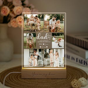 Puede incluir: Un expositor de fotos rectangular de acrílico con base de madera iluminada. La pantalla muestra seis fotografías familiares, el texto "dad", "YOUR STRENGTH INSPIRES ME EVERY DAY", "Matt", "Luna", "Sunny" y "You're my hero" grabado en la base.
