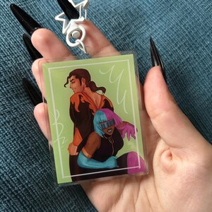 May include: A clear, rectangular acrylic charm featuring a colorful illustration of two figures embracing, set against a light green background. A silver star-shaped ring is visible above the charm. The charm is held in a hand with long, black fingernails.