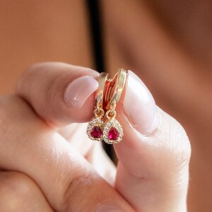 Puede incluir: Un par de pendientes de aro dorados con piedras rojas en forma de lágrima en el centro. Cada piedra está rodeada por un halo de pequeñas piedras claras y brillantes. Los pendientes tienen un mecanismo de cierre de bisagra y acabado dorado.