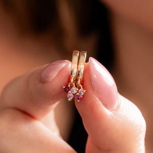 May include: A pair of small gold hoop earrings, each featuring a cluster of three tiny round gemstones in varying shades of pink and deep red. The hoops are polished gold, and the stones are arranged in a triangular formation at the base.