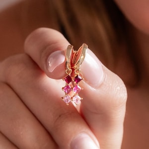 Può includere: Un paio di orecchini a cerchio dorati con un design pendente verticale. Ogni orecchino è decorato con tre pietre preziose a taglio quadrato in una sfumatura di colori, che passa dal rosso intenso in alto al rosa chiaro alla base.