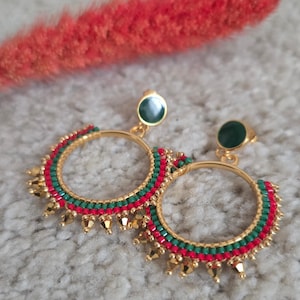Op de afbeelding: Een paar goudkleurige oorringen met rode en groene kralenaccenten. De oorbellen hebben een kleine, donkergroene oorbelknop en goudkleurige, druppelvormige hangers. Een rood, veervormig decoratief element is zichtbaar op de achtergrond.