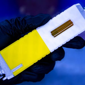 Può includere: Un dispositivo portatile con un corpo giallo e bianco, tenuto da una mano guantata. Il dispositivo ha una forma rettangolare con un componente rettangolare marrone al centro. L'estremità ha un tappo argentato e un pomello nero. Il dispositivo è probabilmente utilizzato per scopi scientifici o medici.