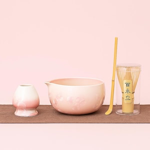 Peut inclure: Un ensemble à thé matcha comprenant un bol en céramique rose avec des motifs floraux, un fouet, une cuillère et un petit récipient en céramique. L'ensemble est disposé sur un tapis marron sur fond rose.