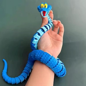 May include: A blue articulated snake toy with a segmented body, featuring a cartoon-style head with large yellow eyes, a red mouth, and white fangs. The snake has a textured, scale-like pattern along its back and is designed to wrap around an object.