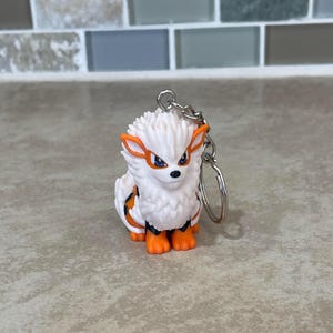 May include: A white and orange Arcanine keychain. The keychain features a small, detailed figurine of the Pok&eacute;mon Arcanine with orange accents on its ears, paws, and markings. The keychain is attached to a silver metal ring.