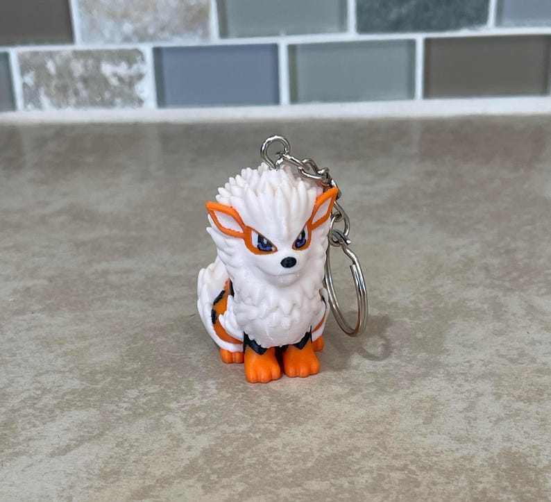 May include: A white and orange Arcanine keychain. The keychain features a small, detailed figurine of the Pok&eacute;mon Arcanine with orange accents on its ears, paws, and markings. The keychain is attached to a silver metal ring.