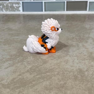 May include: A white and orange figurine of a dog-like creature with spiky fur and orange accents. The figurine has orange goggles and black and orange markings on its back. The creature is in a sitting position.