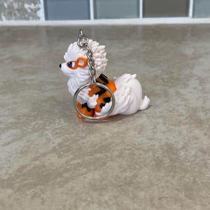 May include: A white and orange dog keychain with a silver keyring. The dog has orange and black markings and orange goggles. The keychain is a small, decorative accessory.