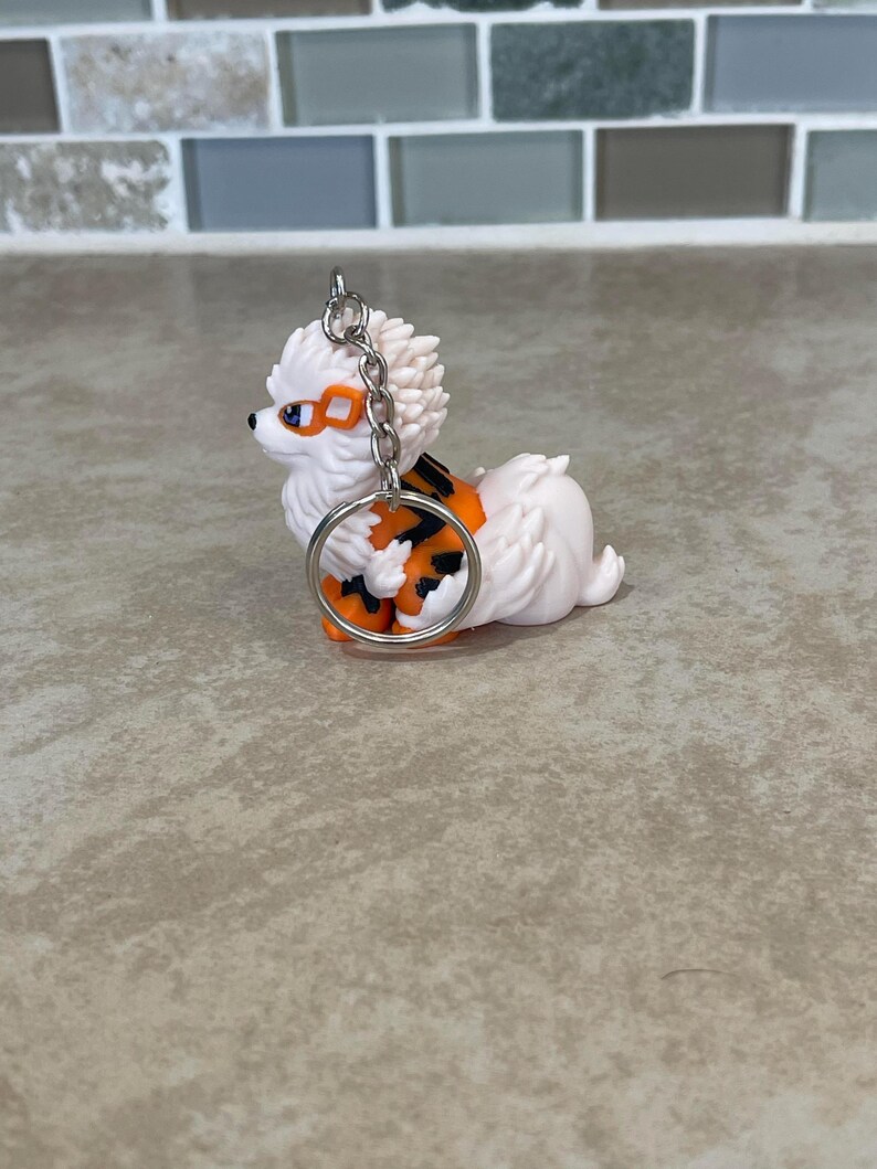 May include: A white and orange dog keychain with a silver keyring. The dog has orange and black markings and orange goggles. The keychain is a small, decorative accessory.