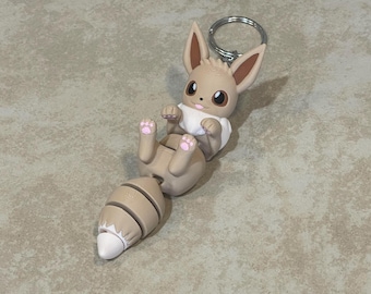 May include: A brown and white Eevee keychain with a silver ring. The Eevee is curled up with its tail wrapped around its body.