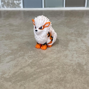 May include: A small, white and orange figurine of a creature with a spiky mane, orange accents, and a black and orange striped pattern on its back. The figurine has orange paws and a determined expression.