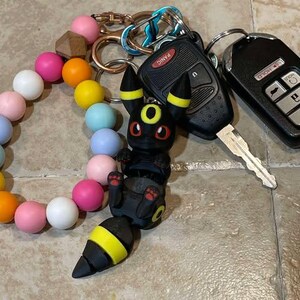 Puede incluir: Un llavero Pokemon negro y amarillo con un ojo rojo y un brazalete de cuentas negro y blanco. El llavero est&aacute; unido a un juego de llaves con un llavero de coche negro que dice "Panic" y una llave plateada.