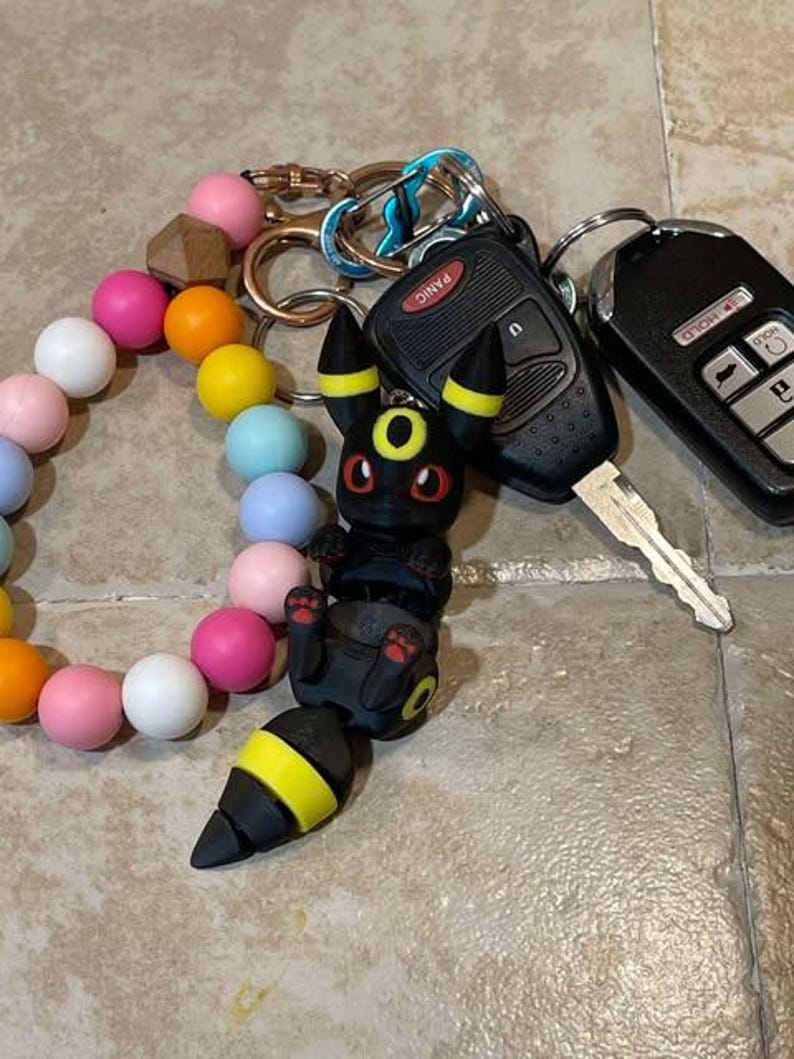 Puede incluir: Un llavero Pokemon negro y amarillo con un ojo rojo y un brazalete de cuentas negro y blanco. El llavero est&aacute; unido a un juego de llaves con un llavero de coche negro que dice "Panic" y una llave plateada.