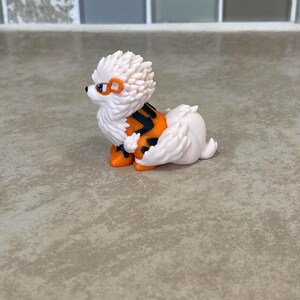 May include: A white and orange figurine of a dog-like creature with a spiky mane, orange accents, and black stripes. The figurine has orange feet and a small tail. The creature has orange goggles and a black nose.