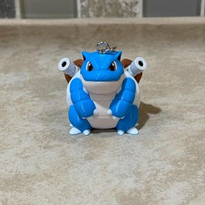 May include: A blue and white 3D printed figurine of a Pokemon character, Squirtle, with a brown shell and two water cannons on its back. The figurine is a keychain.