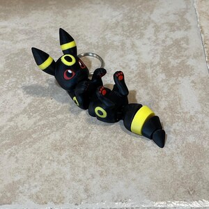 Puede incluir: Llavero Eevee negro y amarillo con ojos rojos. El llavero est&aacute; hecho de pl&aacute;stico y est&aacute; en una pose juguetona.