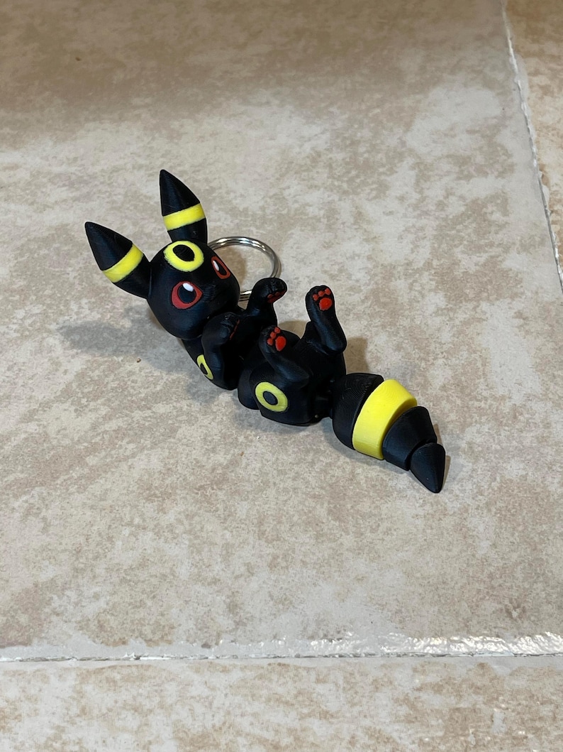 Puede incluir: Llavero Eevee negro y amarillo con ojos rojos. El llavero est&aacute; hecho de pl&aacute;stico y est&aacute; en una pose juguetona.