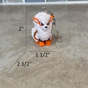 May include: A white and orange Arcanine keychain, measuring approximately 2 inches tall, 2.5 inches long, and 1.5 inches wide. The keychain features a detailed design of the Pok&eacute;mon character with a silver chain and keyring.