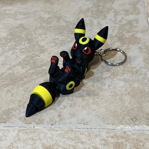 Puede incluir: Llavero Pok&eacute;mon negro y amarillo con una figura 3D del personaje Umbreon. La figura est&aacute; en posici&oacute;n sentada con la cola enroscada alrededor del cuerpo.