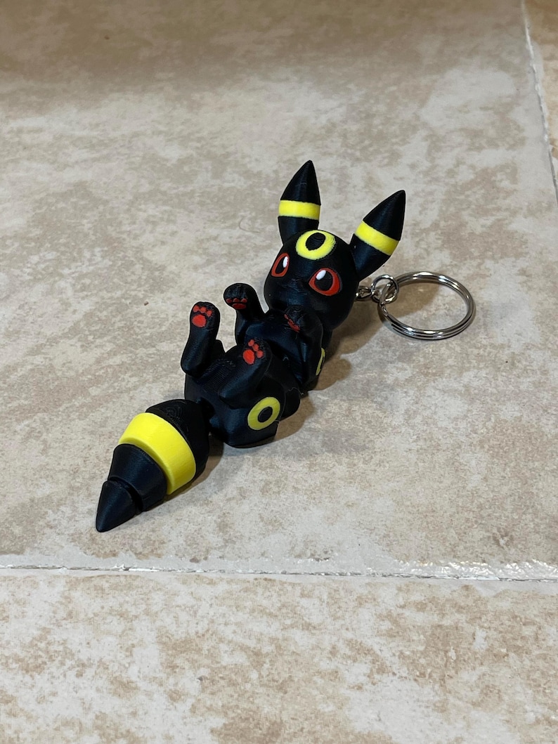 Puede incluir: Llavero Pok&eacute;mon negro y amarillo con una figura 3D del personaje Umbreon. La figura est&aacute; en posici&oacute;n sentada con la cola enroscada alrededor del cuerpo.