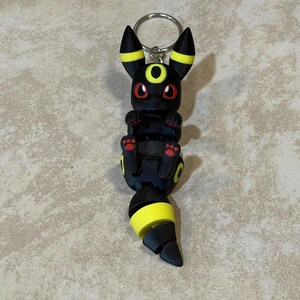Puede incluir: Llavero Pokemon negro y amarillo con una representaci&oacute;n 3D del personaje Umbreon. El llavero est&aacute; hecho de pl&aacute;stico y tiene un anillo de metal plateado.