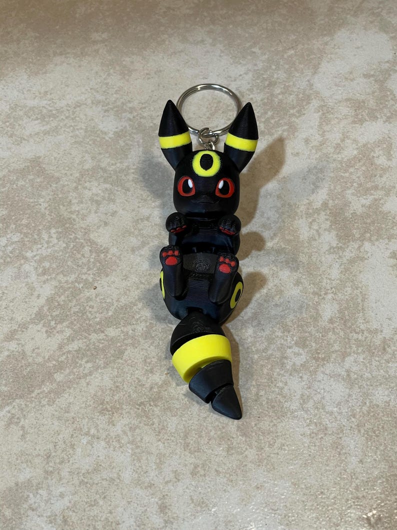 Puede incluir: Llavero Pokemon negro y amarillo con una representaci&oacute;n 3D del personaje Umbreon. El llavero est&aacute; hecho de pl&aacute;stico y tiene un anillo de metal plateado.