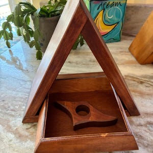 May include: A triangular wooden structure with a dark brown finish, featuring a flat base and a central, removable wooden piece with a circular cutout. The object has a smooth, polished surface and a geometric design, showcasing natural wood grain.