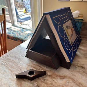 May include: A dark brown wooden book holder with a built-in shelf, made from a repurposed book with blue covers and white lettering. A small, dark brown wooden bookmark lies in front of the book holder.