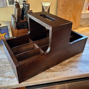 Puede incluir: Un portacubiertos y servilletas de madera marrón oscuro. El organizador tiene múltiples compartimentos para cubiertos y una ranura superior para servilletas. La veta de la madera es visible y el acabado es liso. Diseñado para cocina o comedor.