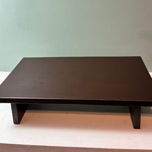 May include: A rectangular wooden riser with a dark brown finish. The platform features a smooth, flat top surface supported by two vertical wooden legs positioned at each end, creating a low-profile stand for elevated display purposes.