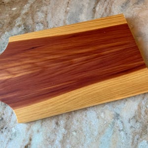 May include: A rectangular wooden chopping board with a curved edge. The centre is a rich reddish-brown, framed by a lighter, golden-toned wood. The board is made of two different types of wood, creating a striking contrast.