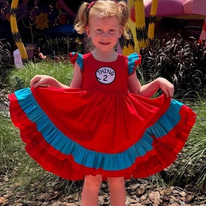 May include: A young girl wearing a red dress with a blue ruffle and a white circle with the text "Thing 2" on it. She is standing in front of a colorful, whimsical background.