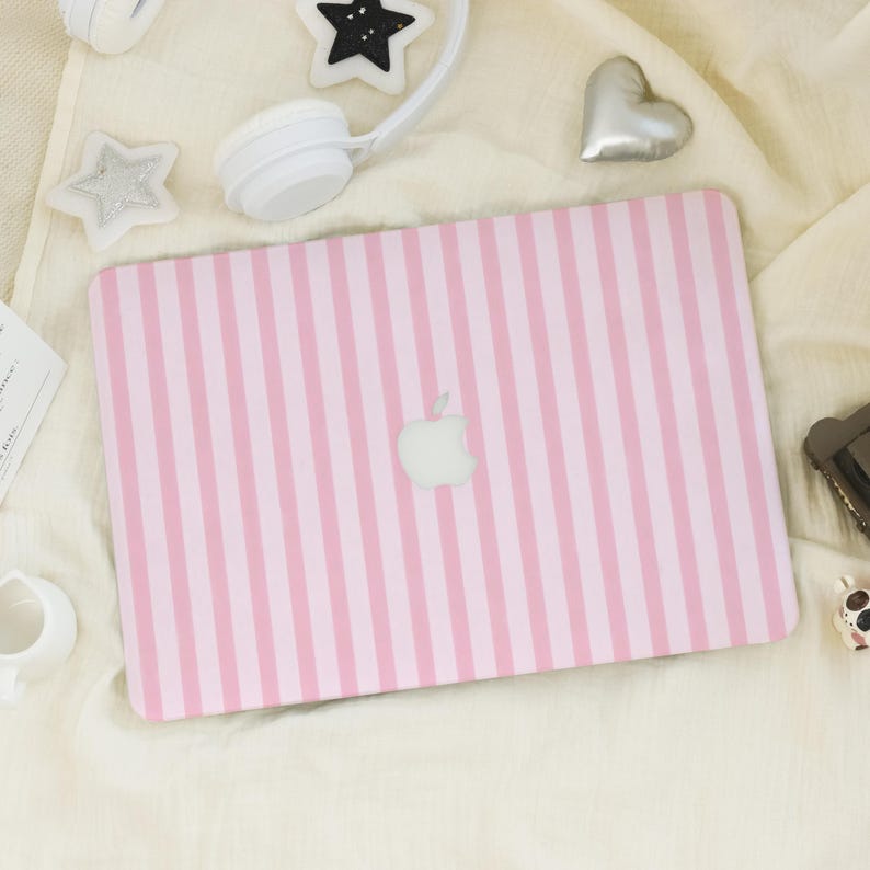 K&ouml;nnte beinhalten: Eine rosa und hellrosa gestreifte Laptop-H&uuml;lle mit dem Apple-Logo in der Mitte. Die H&uuml;lle ist von wei&szlig;en Kopfh&ouml;rern, sternf&ouml;rmigen Dekorationen und einem silbernen Herz auf einer wei&szlig;en Oberfl&auml;che umgeben.