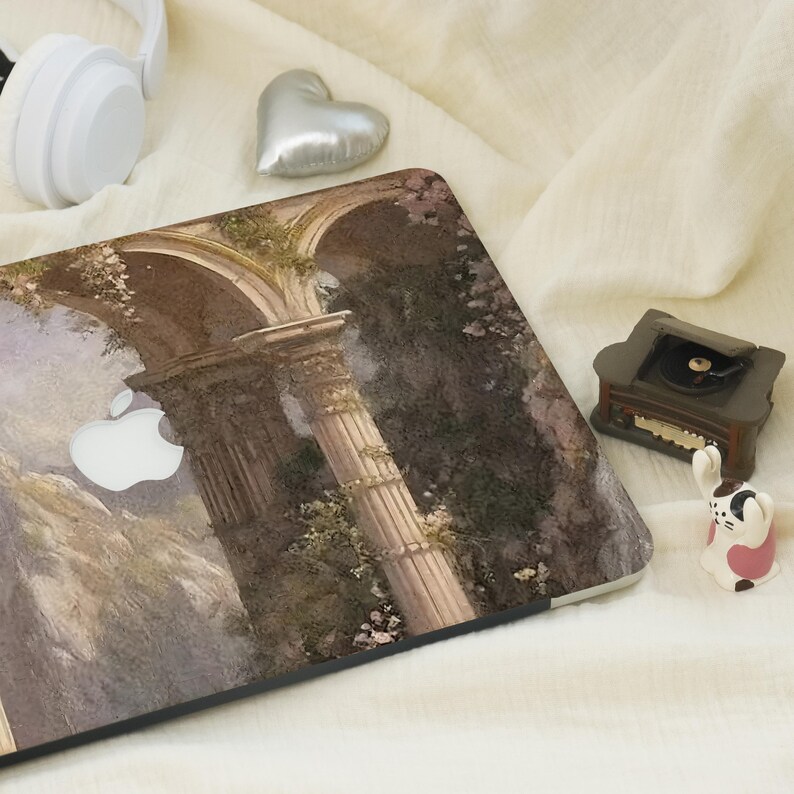 May include: A laptop case featuring a classical architectural design with columns and arches, in shades of beige, green, and brown. A white Apple logo is visible. The case is surrounded by a white headset, a silver heart-shaped decoration, and a miniature record player.