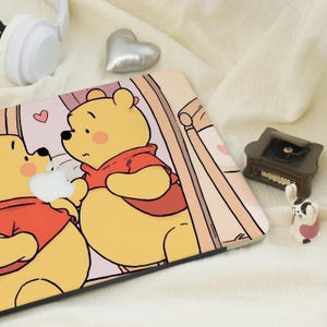 May include: A laptop skin featuring a cartoon illustration of Winnie the Pooh and a friend, both in red tops. The skin has a light pink background with a heart and a window design. A small record player and a rabbit figurine are also visible.